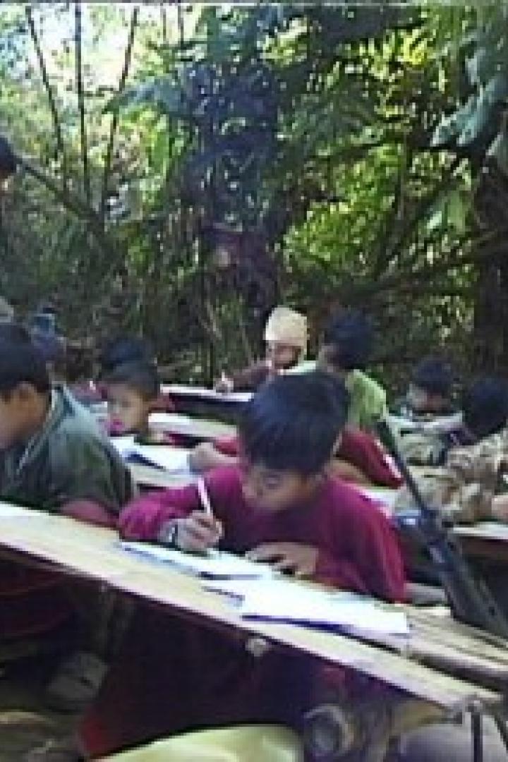 Kinder sitzen an Tischen im Freien und schreiben, im Hintergrund ein bewaffneter Mann.