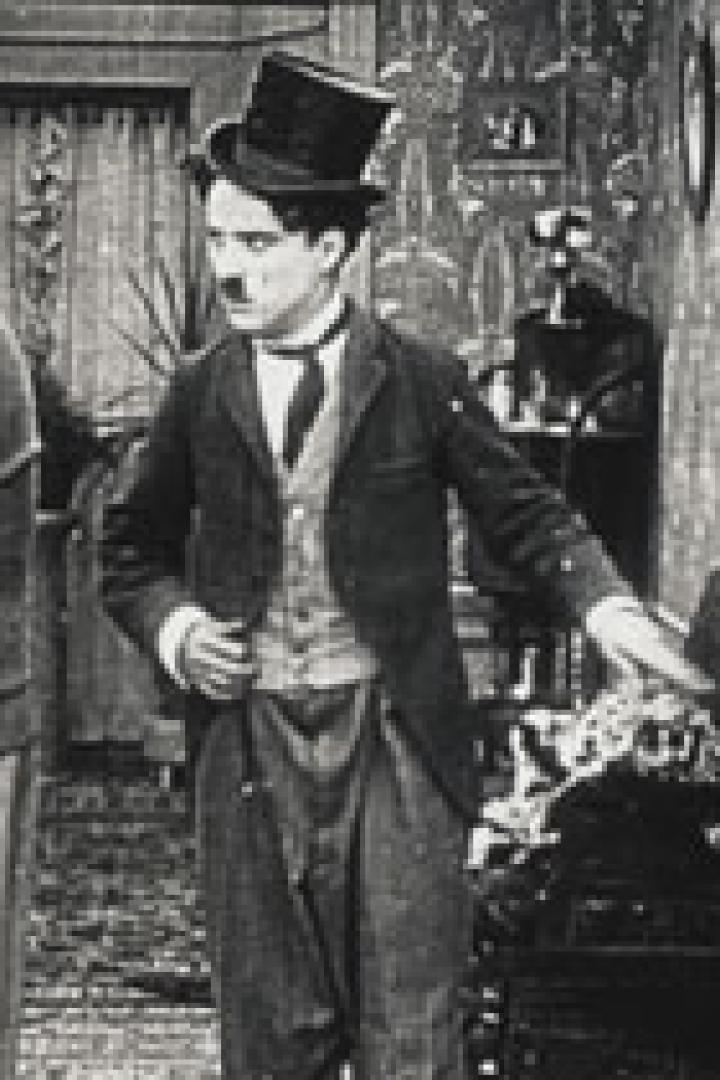 Charlie Chaplin in Anzug und Melone gestikuliert in einem Raum, in dem eine weitere Person steht.