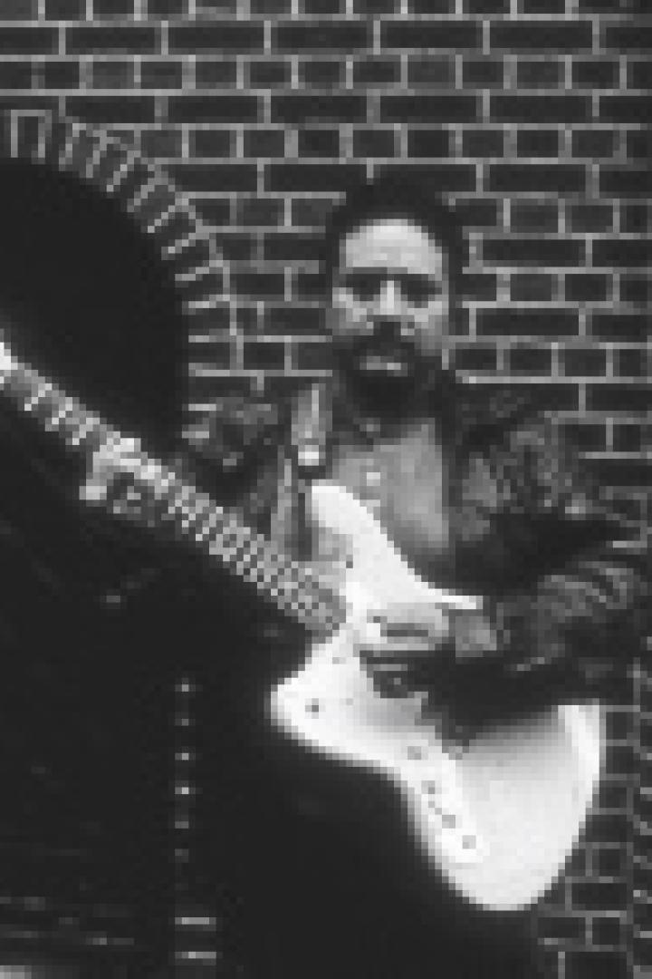 Ein Mann mit Gitarre posiert vor einer Backsteinmauer.
