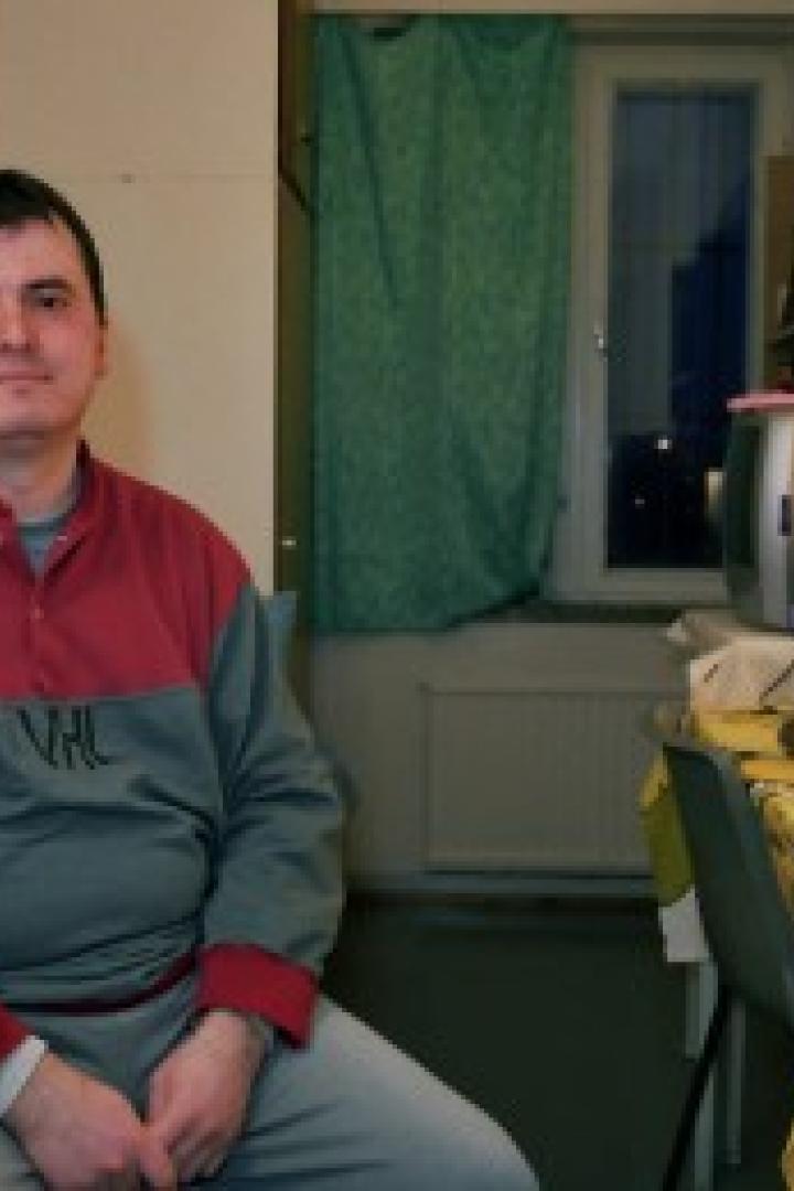 Ein Mann sitzt in einem Zimmer mit Schreibtisch und einem kleinen Fernseher.