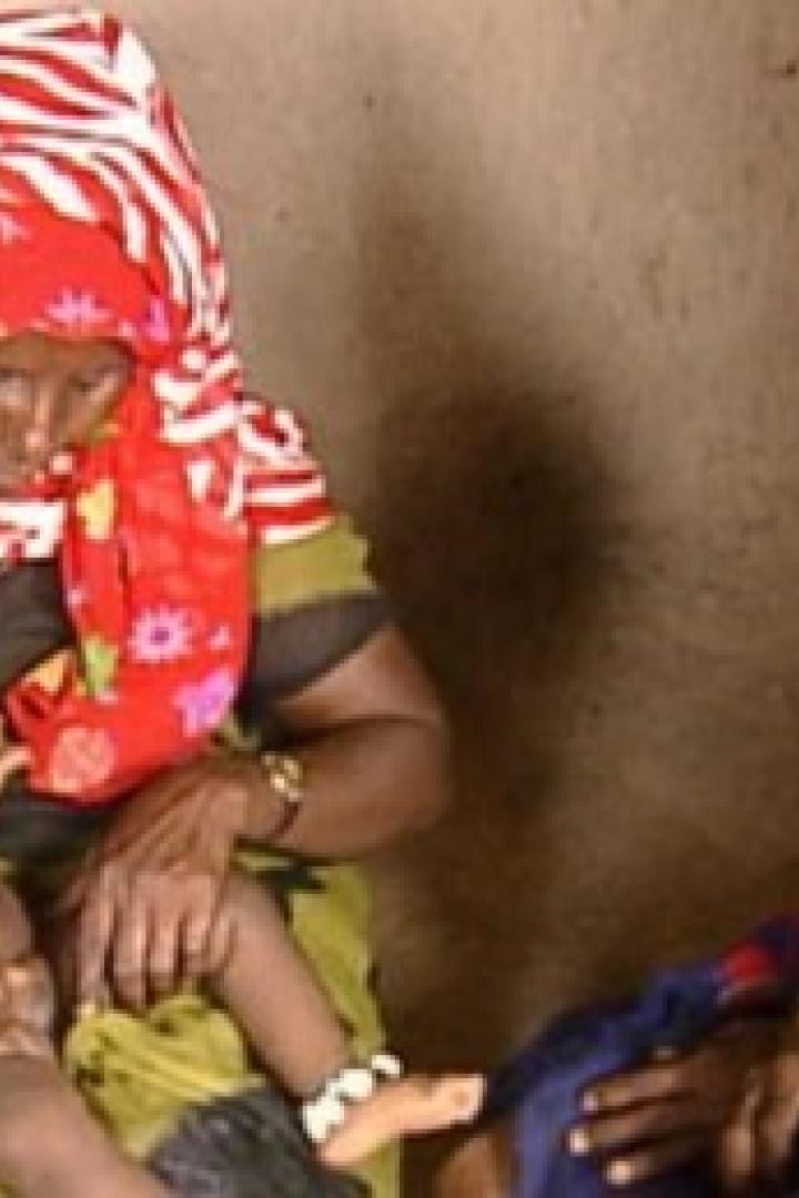 Eine Frau mit einem rot-weiß gemusterten Kopftuch hält ein Baby im Arm, während eine andere Frau zusieht.