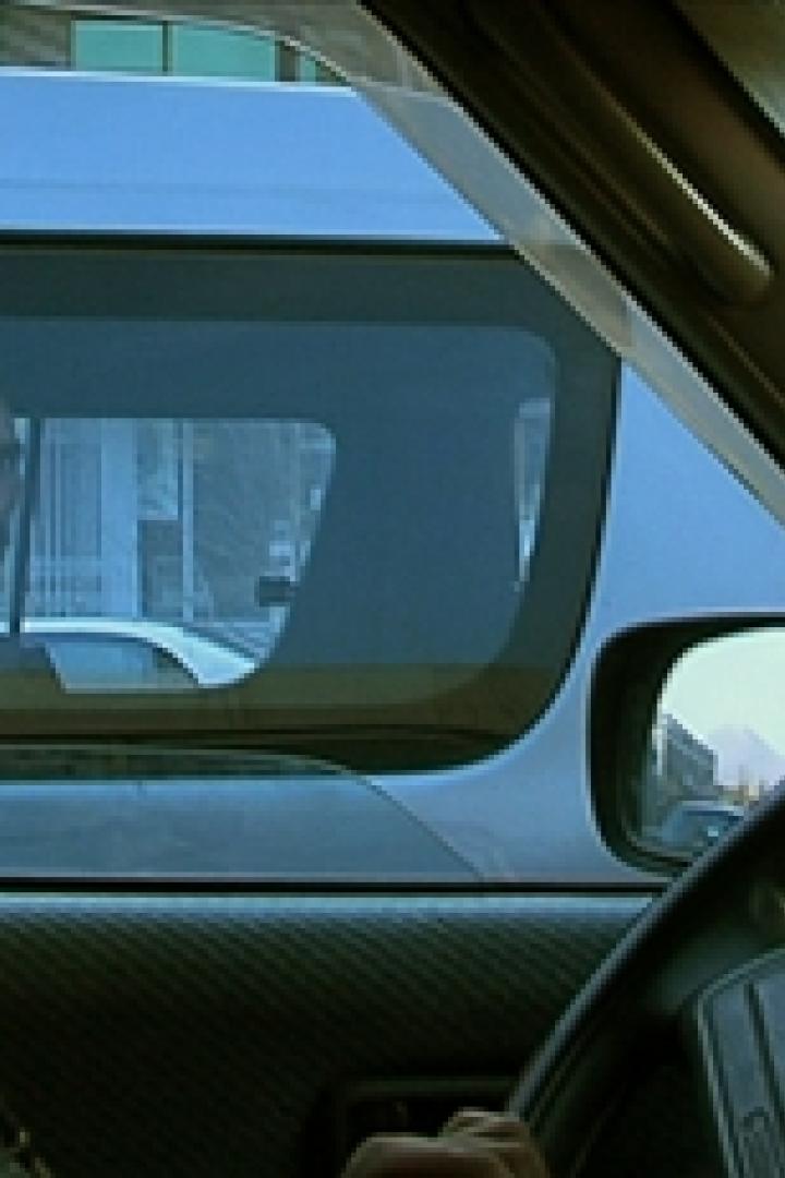 Eine Frau mit Brille fährt ein Auto; im Hintergrund ist ein Mann im Auto hinter ihr zu sehen.