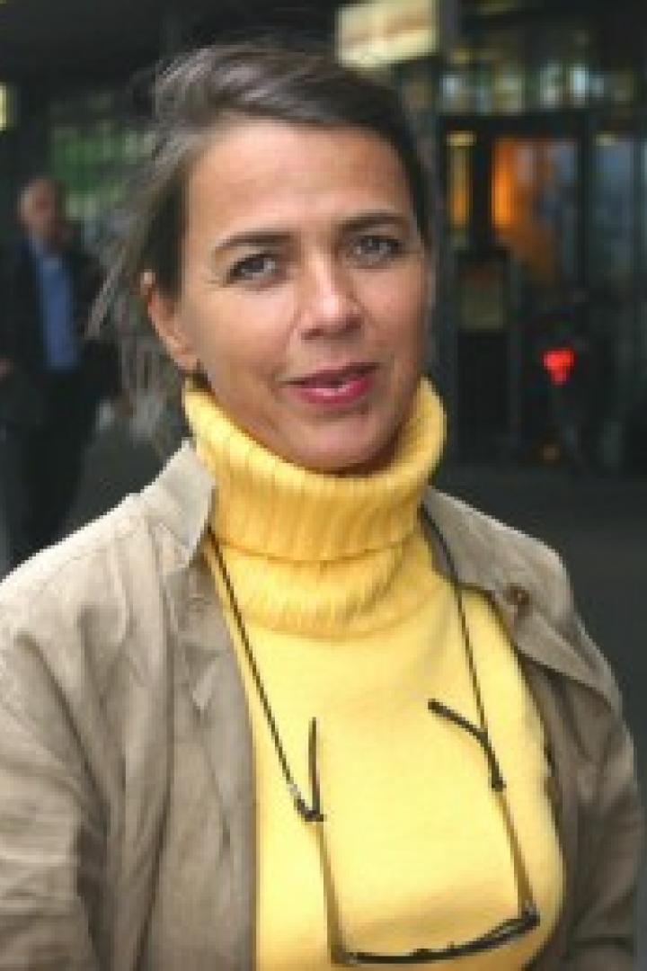 Eine Frau mit gelbem Rollkragenpullover und Brille steht auf einem Bürgersteig.