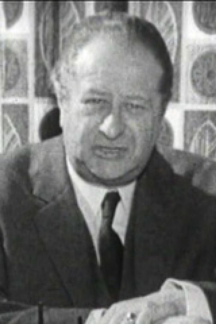 Ein Mann im Anzug sitzt vor einem gemusterten Vorhang.