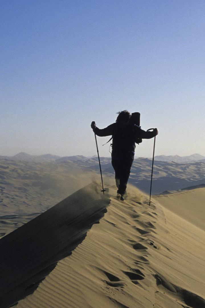 Ein Wanderer mit Rucksack und Stöcken geht über eine Sanddüne.