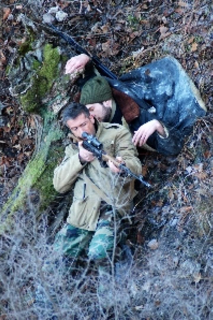 Zwei Männer liegen im Unterholz, einer zielt mit einem Gewehr.