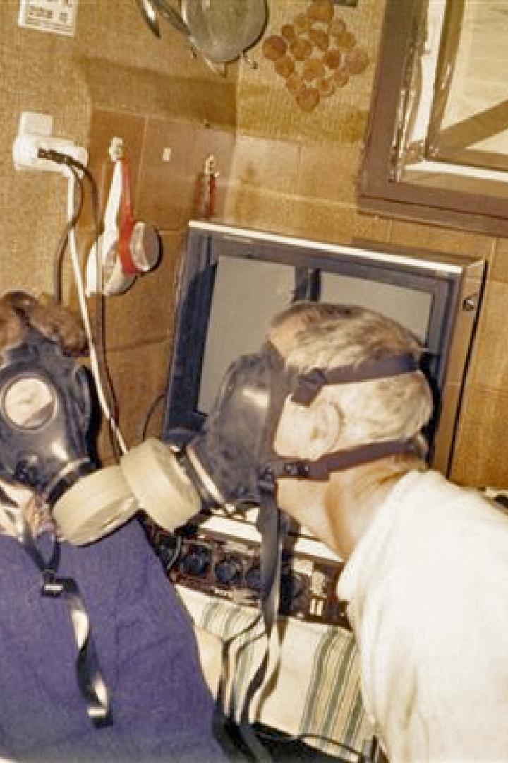 Zwei Personen mit Gasmasken stehen vor einem alten Fernseher.