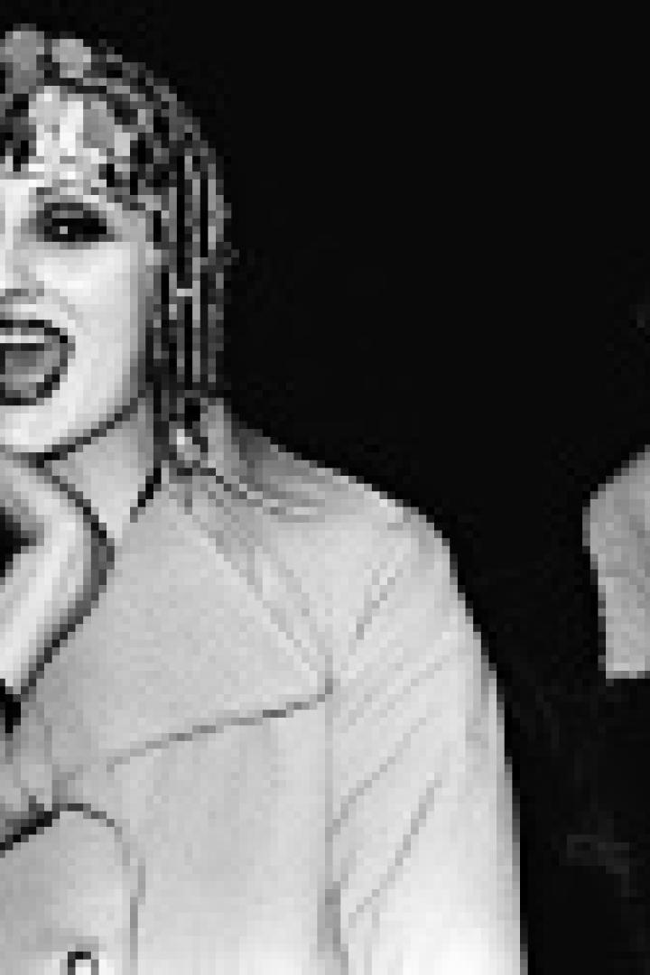 Eine Frau mit Kopfschmuck und Trenchcoat steht neben Andy Warhol.