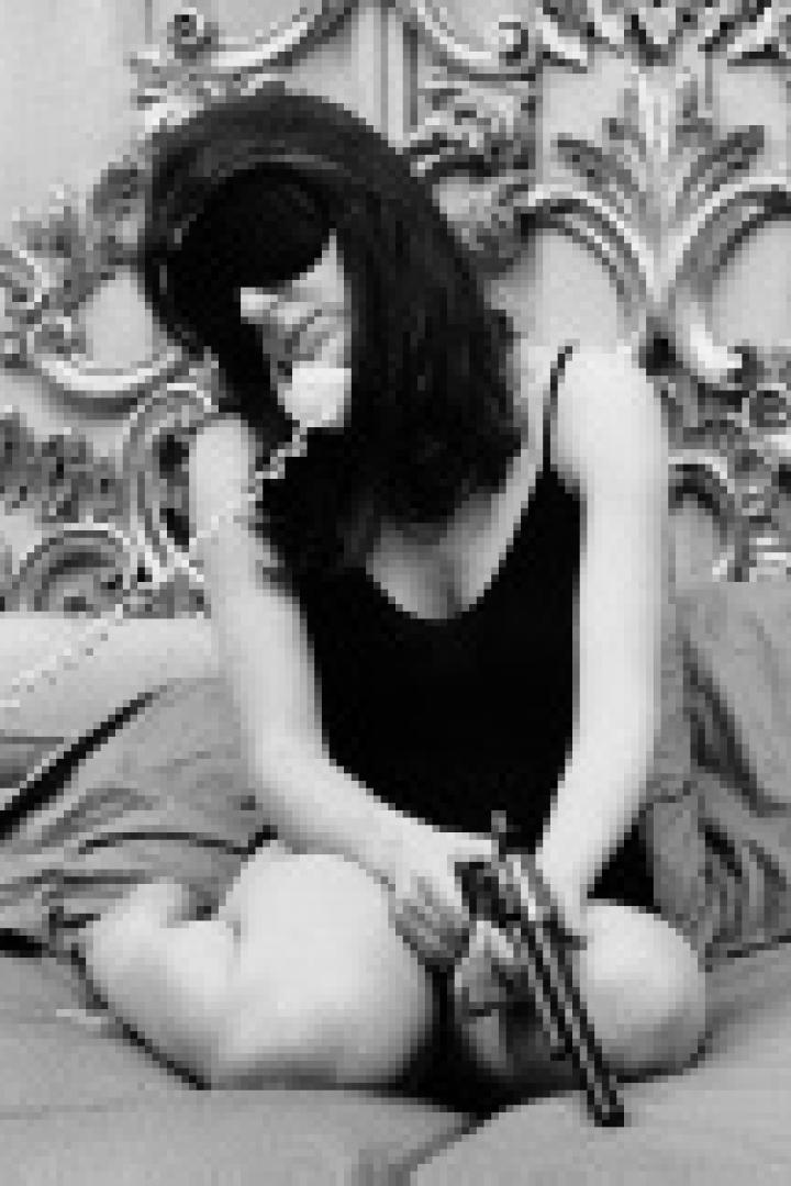 Eine Frau sitzt mit verbundenen Augen auf einem Bett, telefoniert und hält eine Pistole in der Hand.