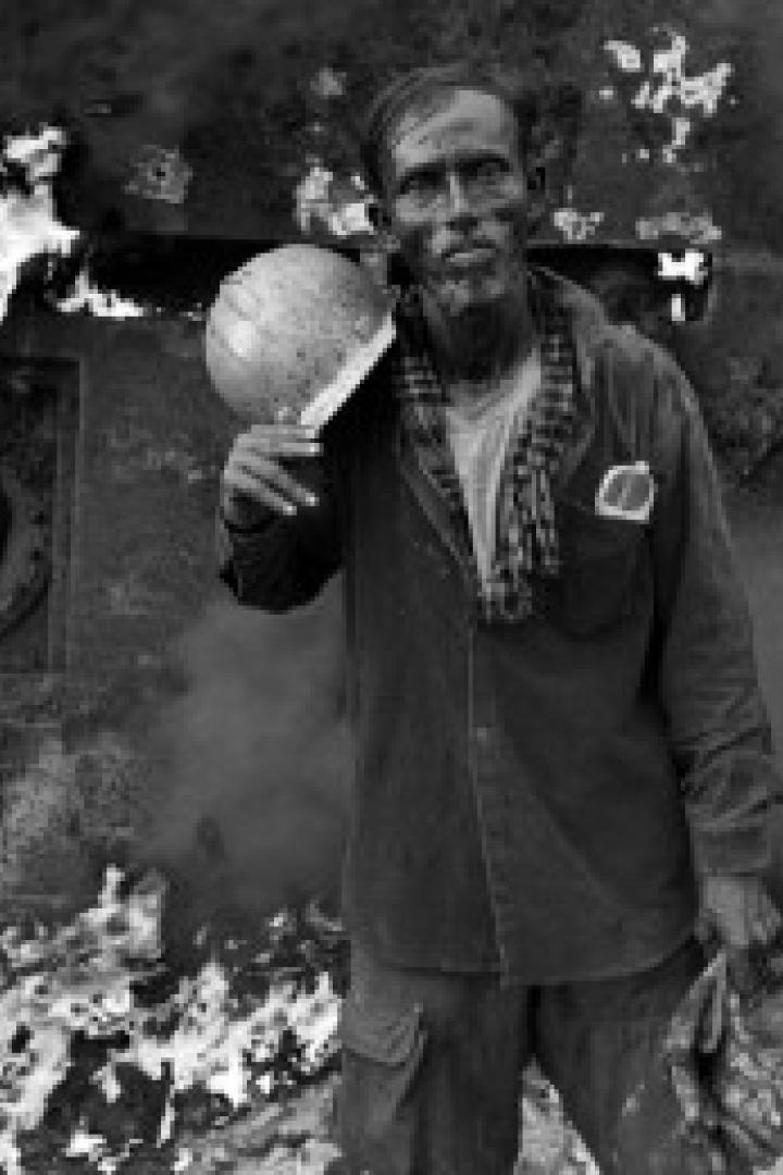 Ein erschöpfter Arbeiter hält einen Helm vor einem zerstörten, rauchenden Gebäude.