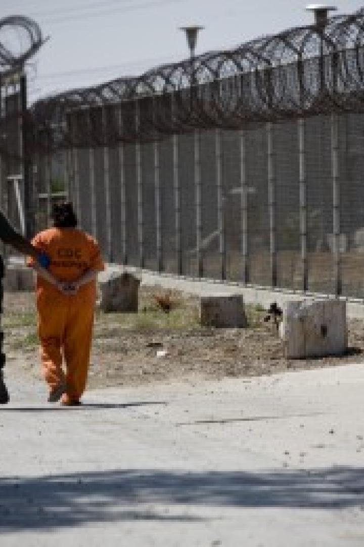 Ein Gefängniswärter begleitet einen Gefangenen in oranger Kleidung entlang eines Zauns mit Stacheldraht.