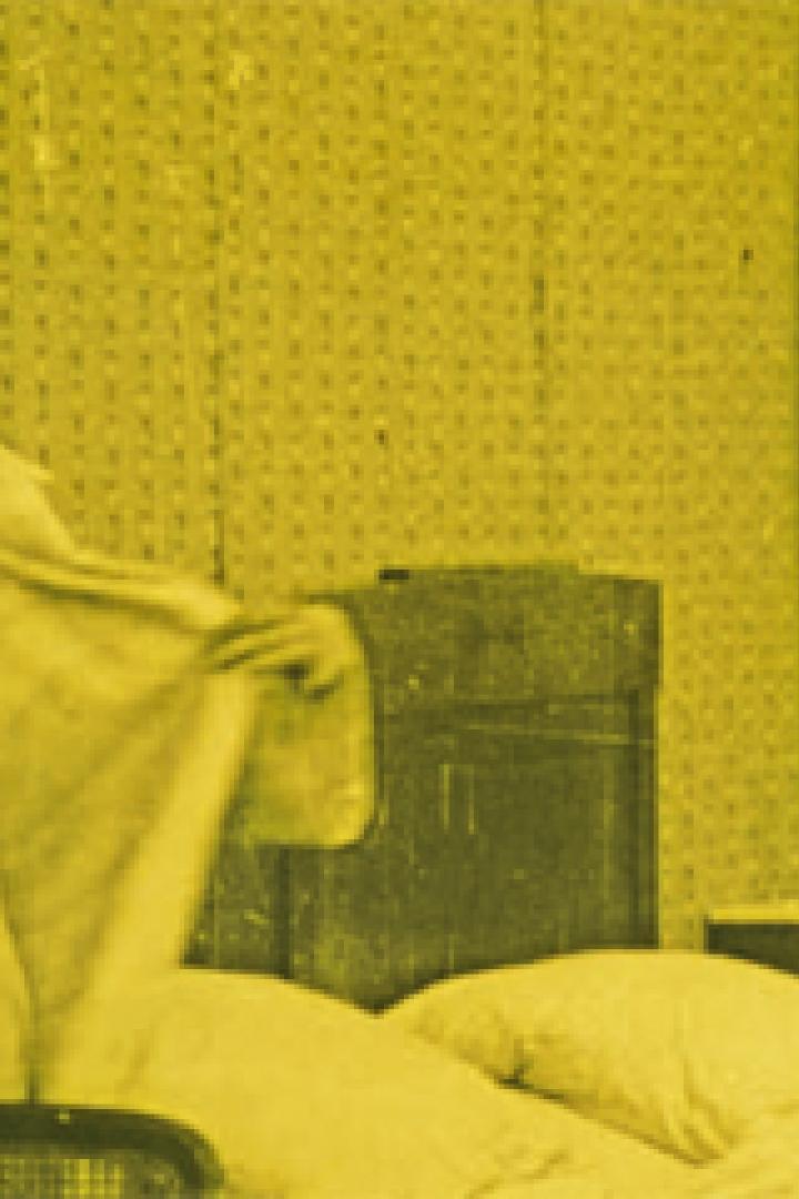 Eine Frau im Nachthemd steht lächelnd neben einem Bett.