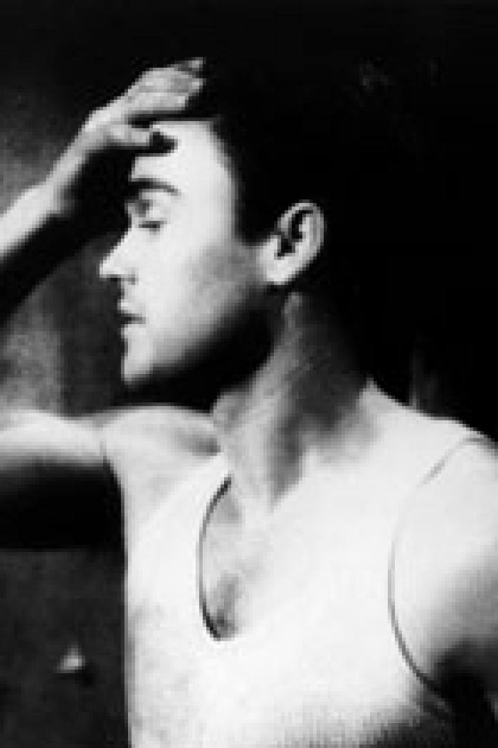 Ein Mann mit Tätowierung und Unterhemd hält sich die Hand an die Stirn.