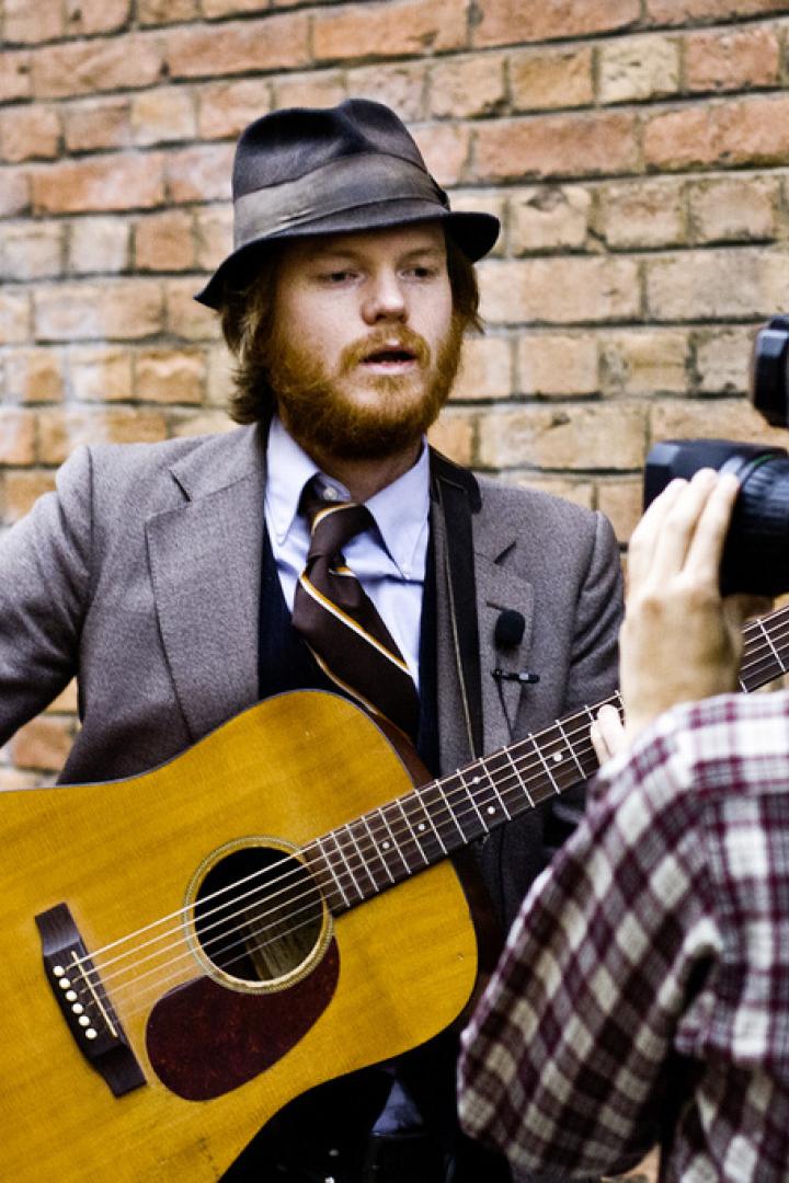 Ein Musiker mit Hut und Gitarre wird von einem Kameramann gefilmt.