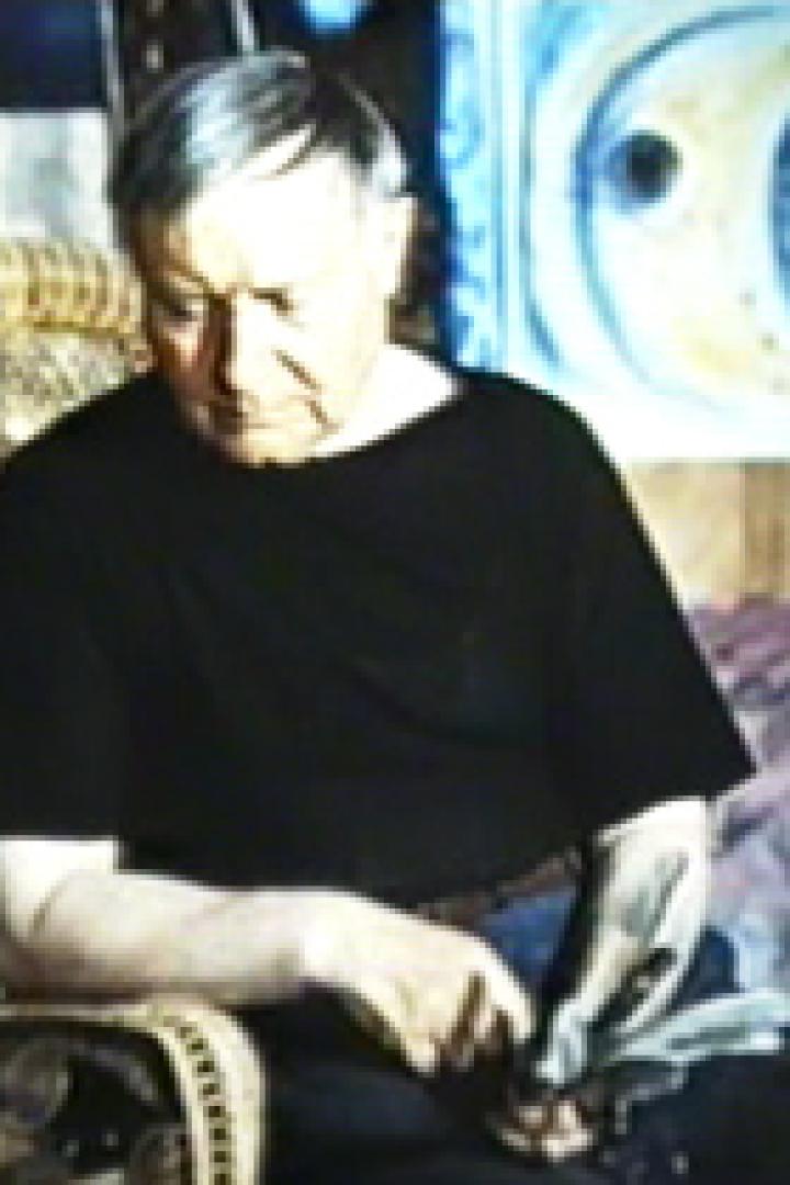 Ein älterer Mann sitzt in einem Korbstuhl vor einem abstrakten Gemälde.