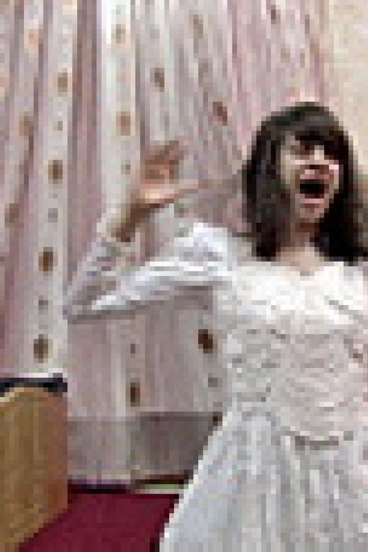 Eine Person im weißen Kleid steht mit erhobenen Händen und geöffnetem Mund in einem Raum.