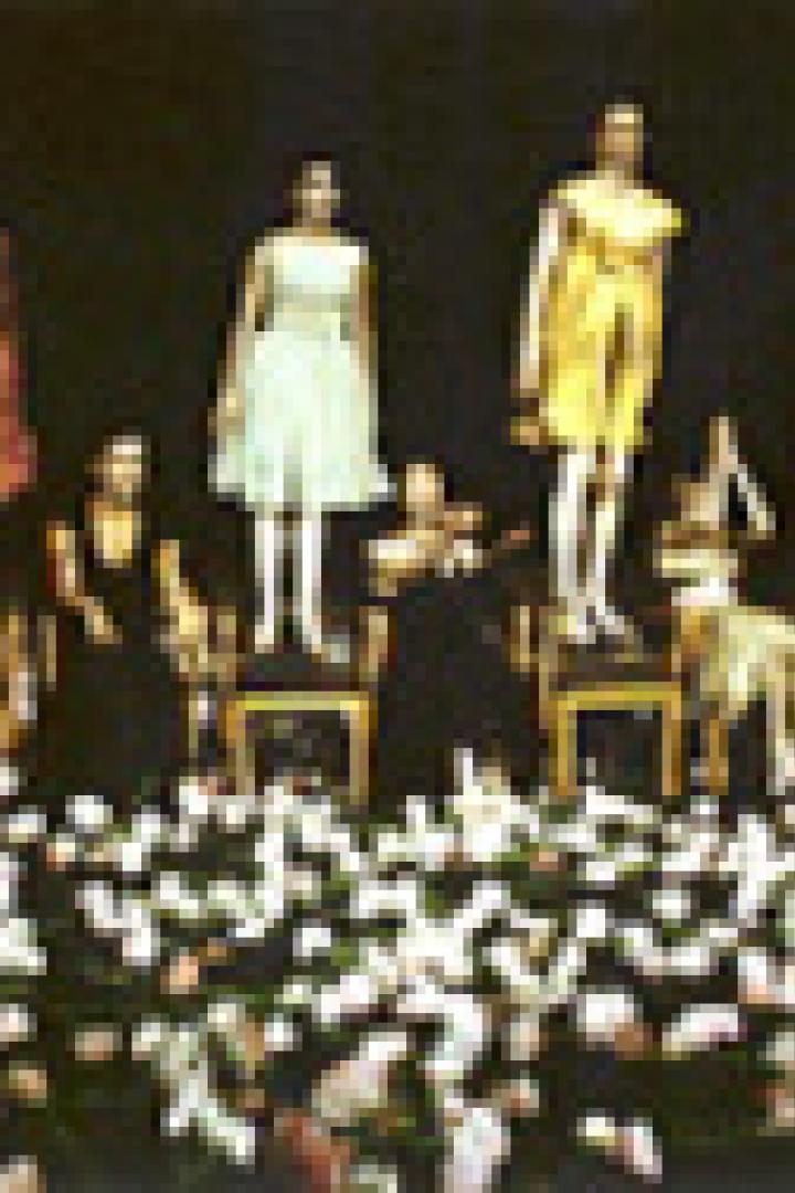 Eine Modenschau mit Models in farbenfrohen Kleidern, die auf Stühlen vor einem großen Publikum sitzen.