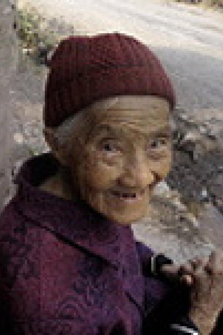 Eine ältere Frau mit roter Mütze sitzt an einer Wand und lächelt.