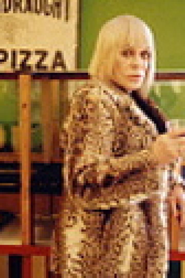 Eine Frau mit blonden Haaren und einem gemusterten Mantel hält ein Glas in der Hand.