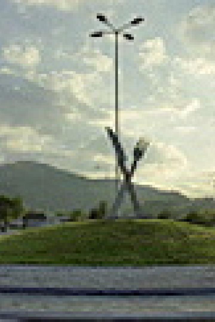 Ein Kreisverkehr mit einer Skulptur aus Gabeln und einem hohen Mast mit Lampen vor einer Bergkulisse.