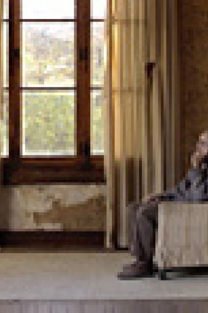 Ein Mann steht am Fenster, während ein älterer Mann in einem Sessel sitzt.