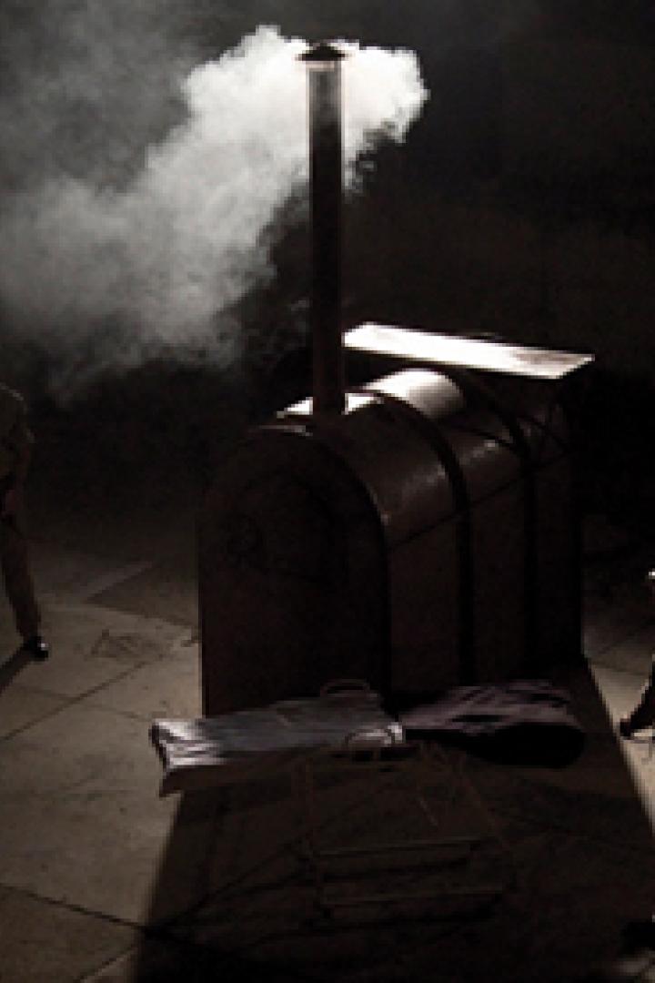 Zwei uniformierte Männer stehen im Dunkeln neben einem Ofen, aus dem Rauch aufsteigt.