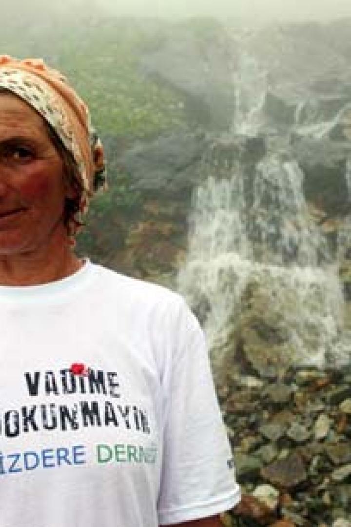 Eine Frau mit Kopftuch und einem T-Shirt mit der Aufschrift „VADIME DOKUNMAYIN“ vor einem Wasserfall.