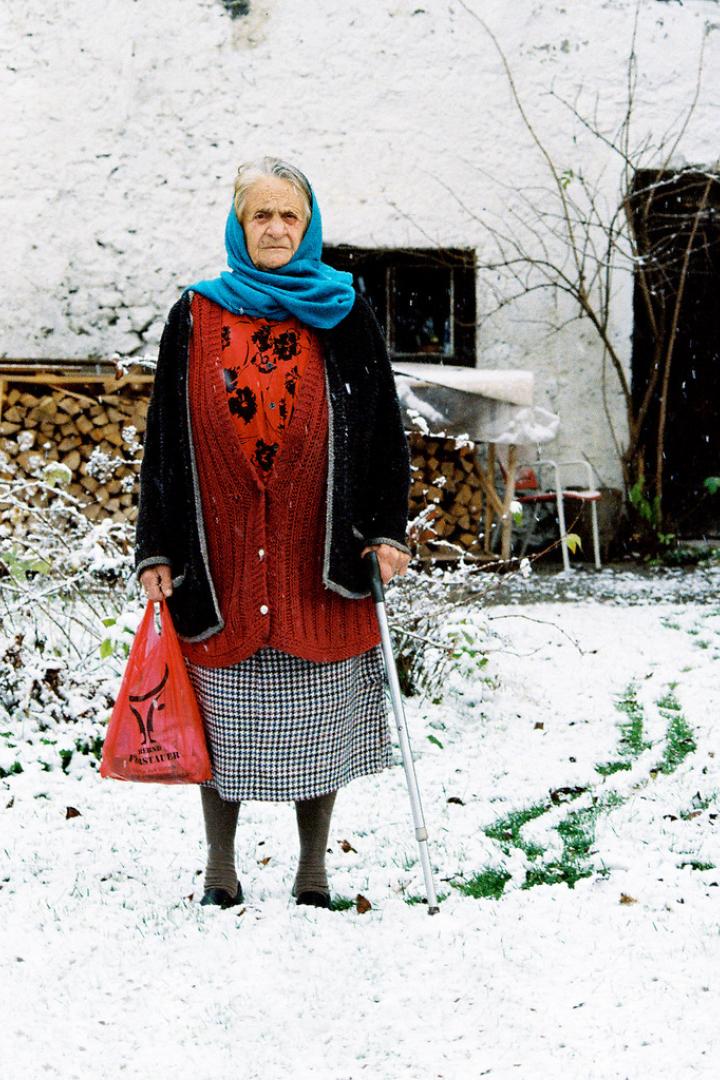 Eine ältere Frau mit Kopftuch und Stock steht im Schnee vor einem Haus.