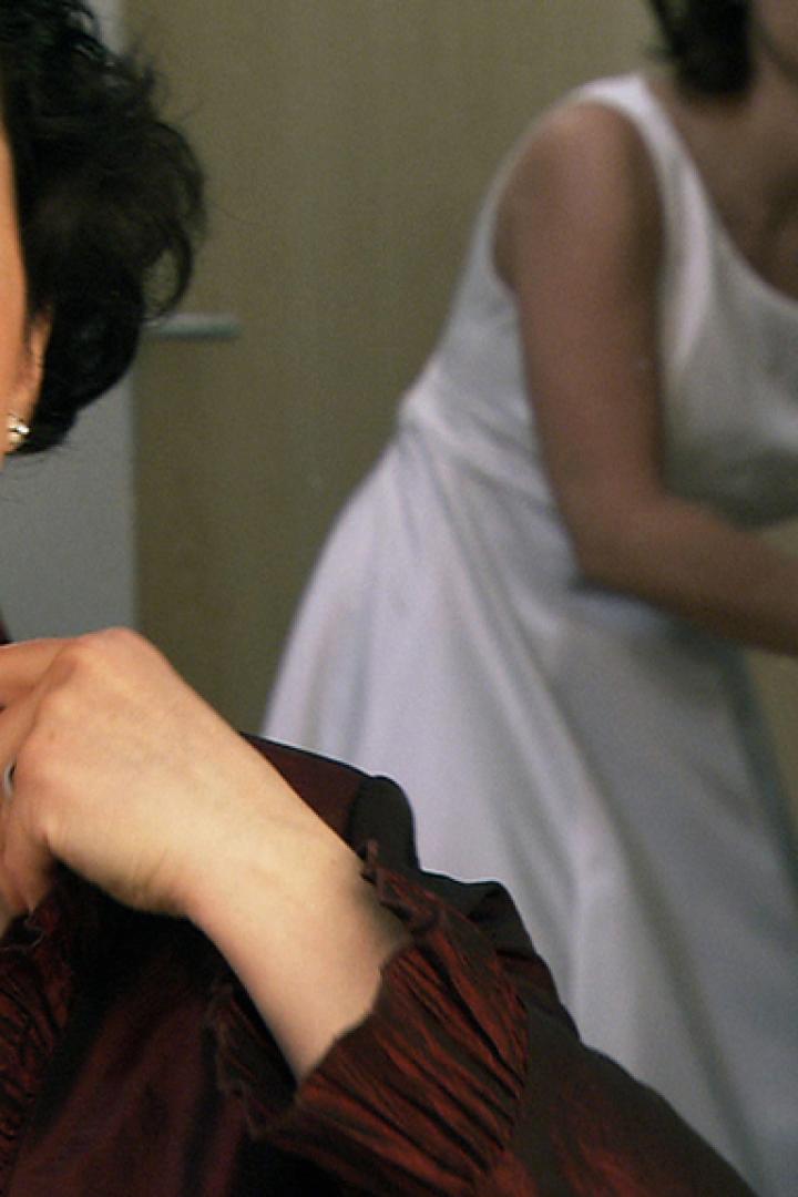 Eine Frau legt eine Halskette an, während eine andere Frau im Hintergrund ein weißes Kleid trägt.