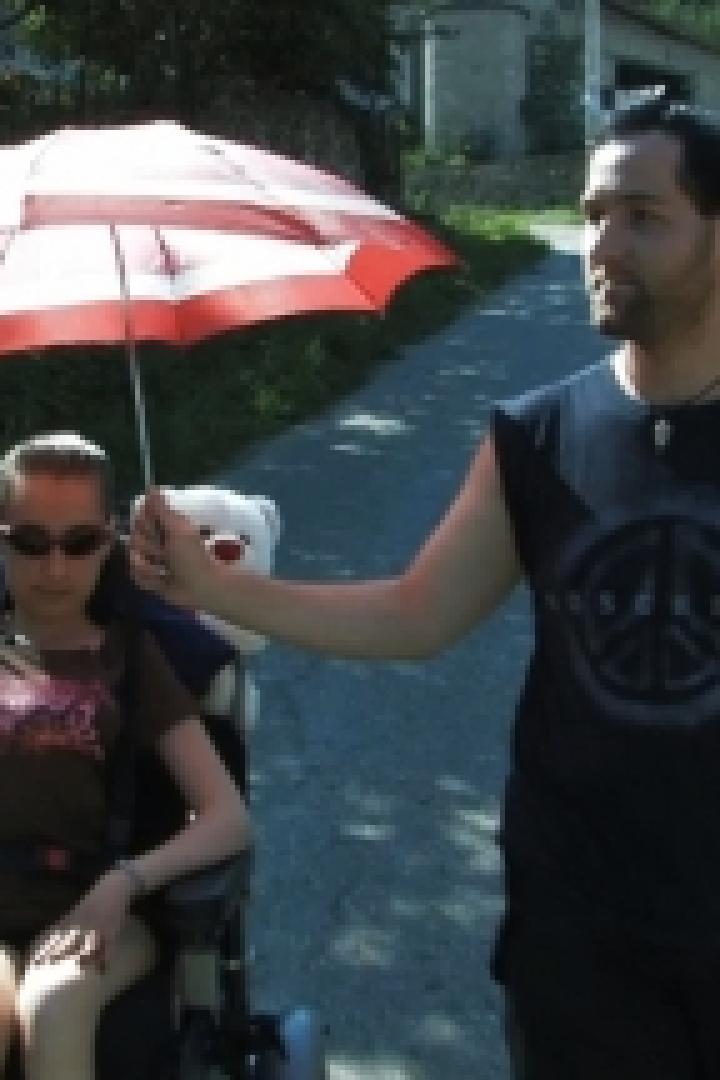 Ein Mann schützt eine Frau im Rollstuhl mit einem rot-weißen Sonnenschirm vor der Sonne.