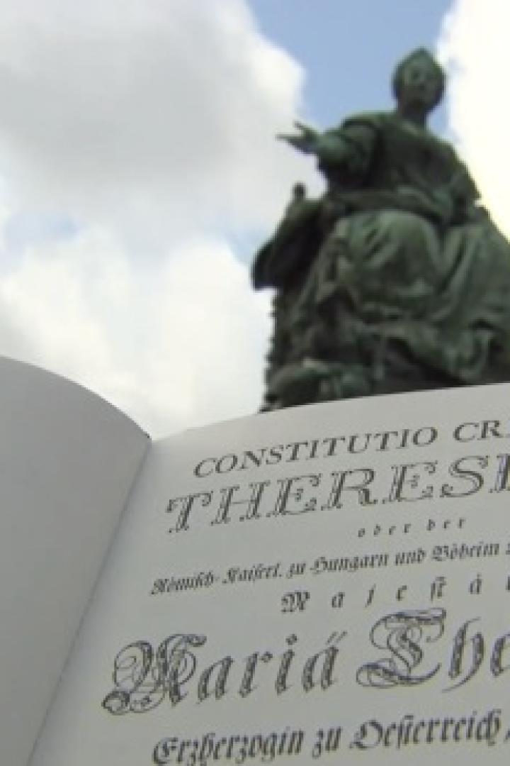 Ein aufgeschlagenes Buch mit dem Titel „Constitutio Criminalis Theresiana“ vor einem Denkmal von Maria Theresia.