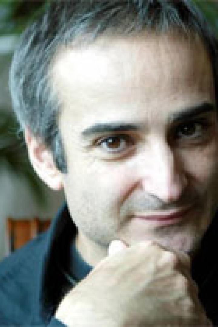 Ein Mann mit dunklen Haaren und einem schwarzen Hemd posiert mit seiner Hand am Kinn.