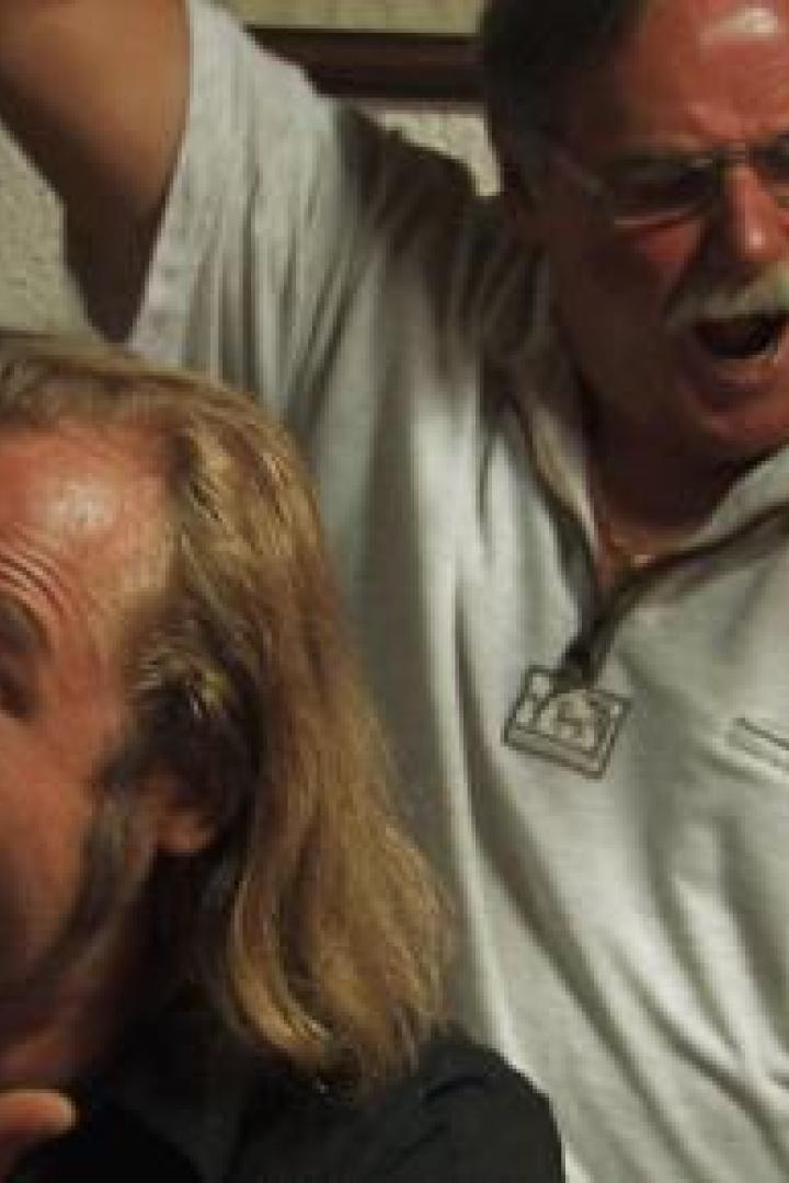 Zwei Männer, einer mit langen blonden Haaren und einer mit Brille, scheinen ausgelassen zu feiern.