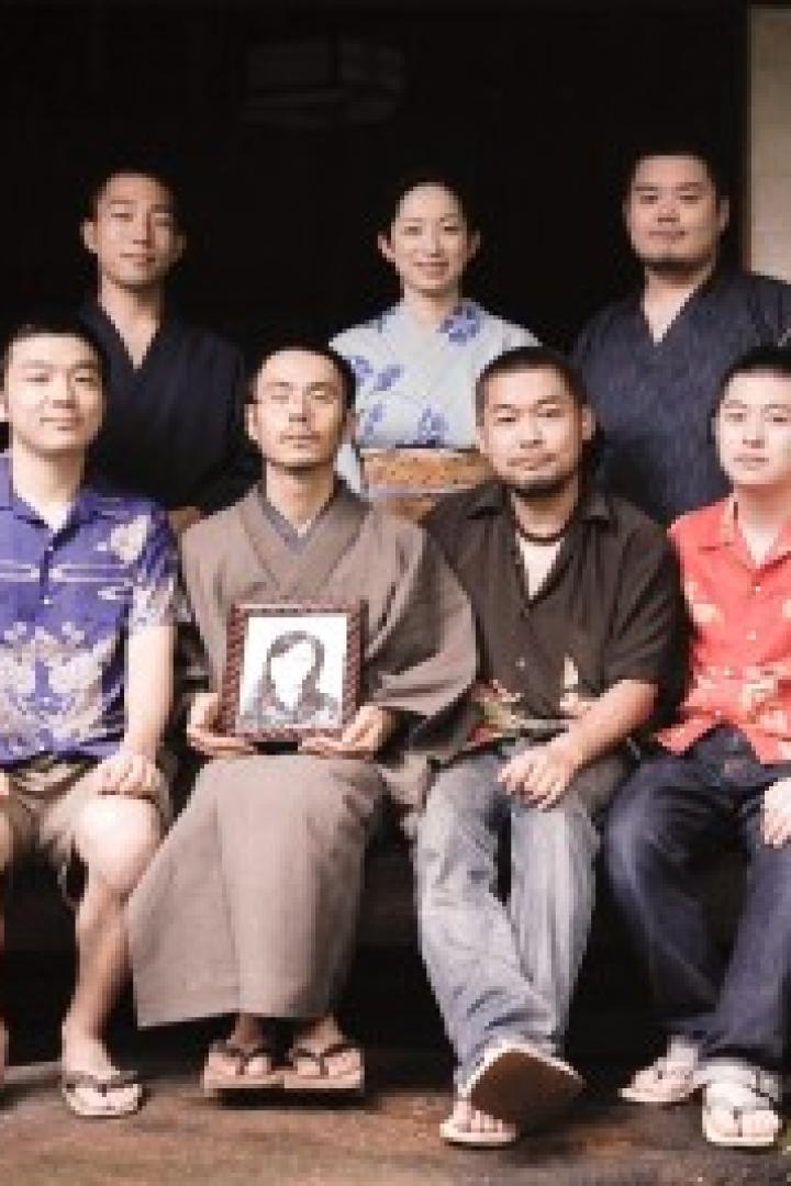 Eine Gruppe von sieben Personen posiert vor einem traditionellen japanischen Haus, eine Person hält ein Porträt.