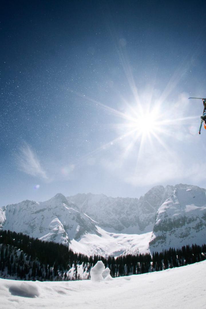 Ein Skifahrer macht einen Rückwärtssalto in der Luft vor einer verschneiten Berglandschaft.