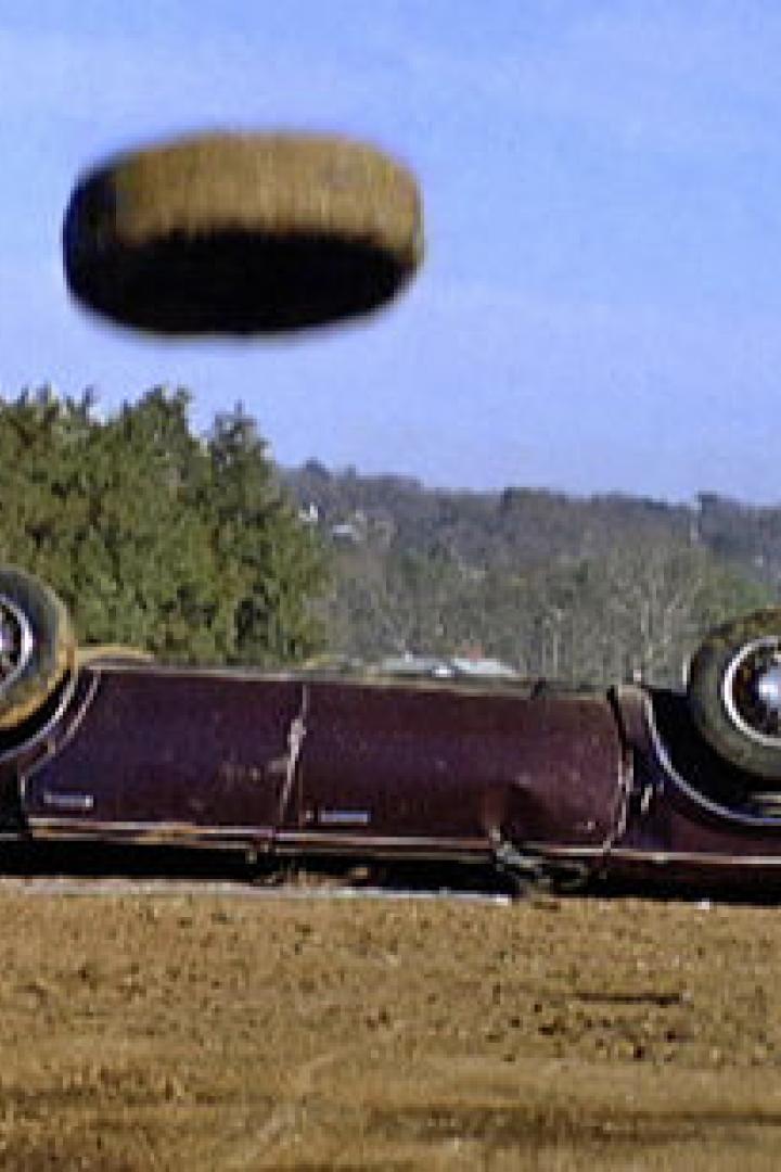Ein umgedrehtes, beschädigtes Auto auf einem Schrottplatz mit einem fliegenden Reifen.