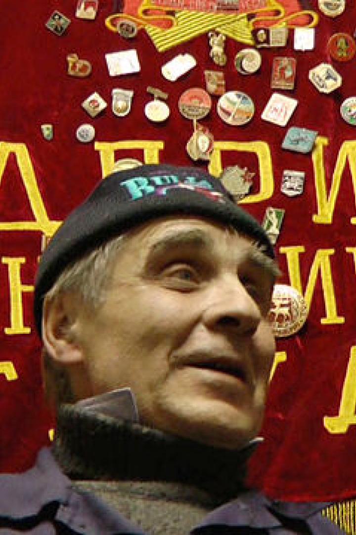 Ein Mann mit Mütze vor einer roten Fahne mit kyrillischer Schrift und Abzeichen.