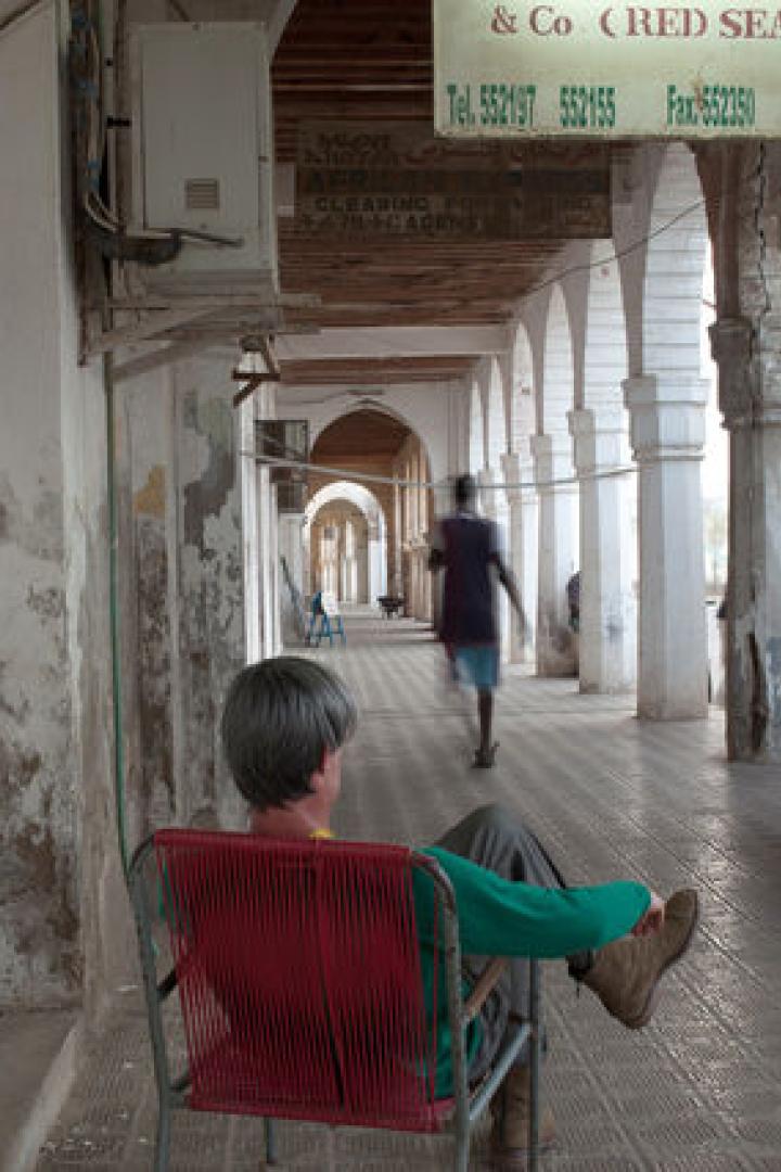 Ein Mann sitzt in einem roten Stuhl unter einem Säulengang, während eine Person vorbeigeht.