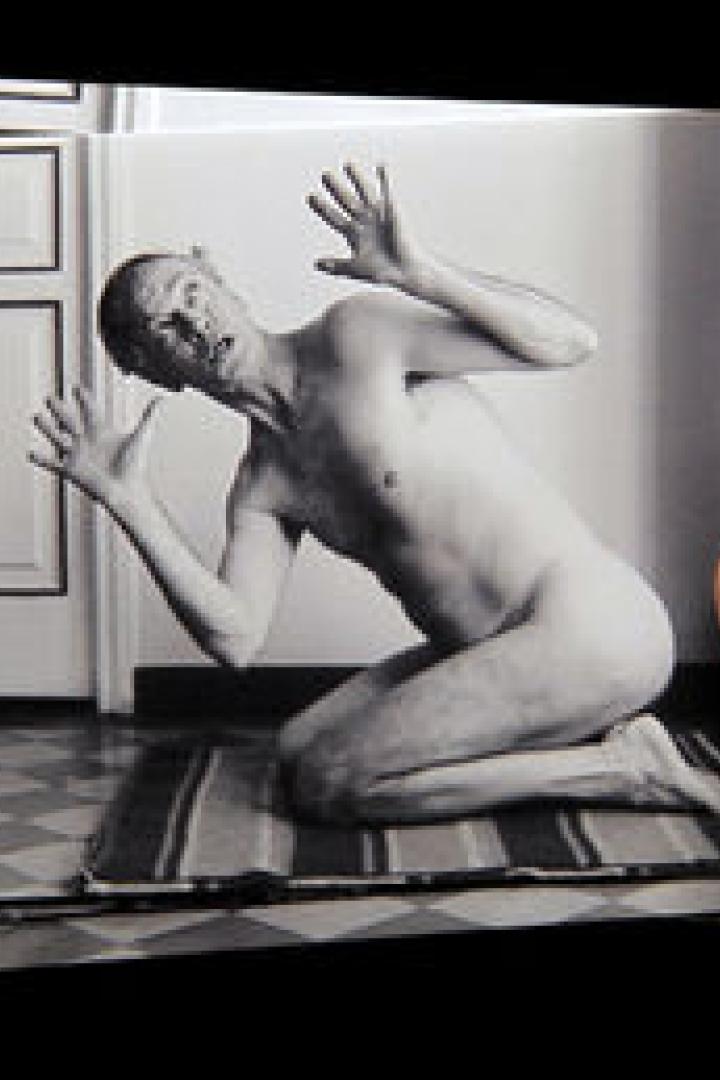 Eine Hand hält ein Schwarzweißfoto eines nackten Mannes, der auf einem Teppich kniet und die Hände hebt.