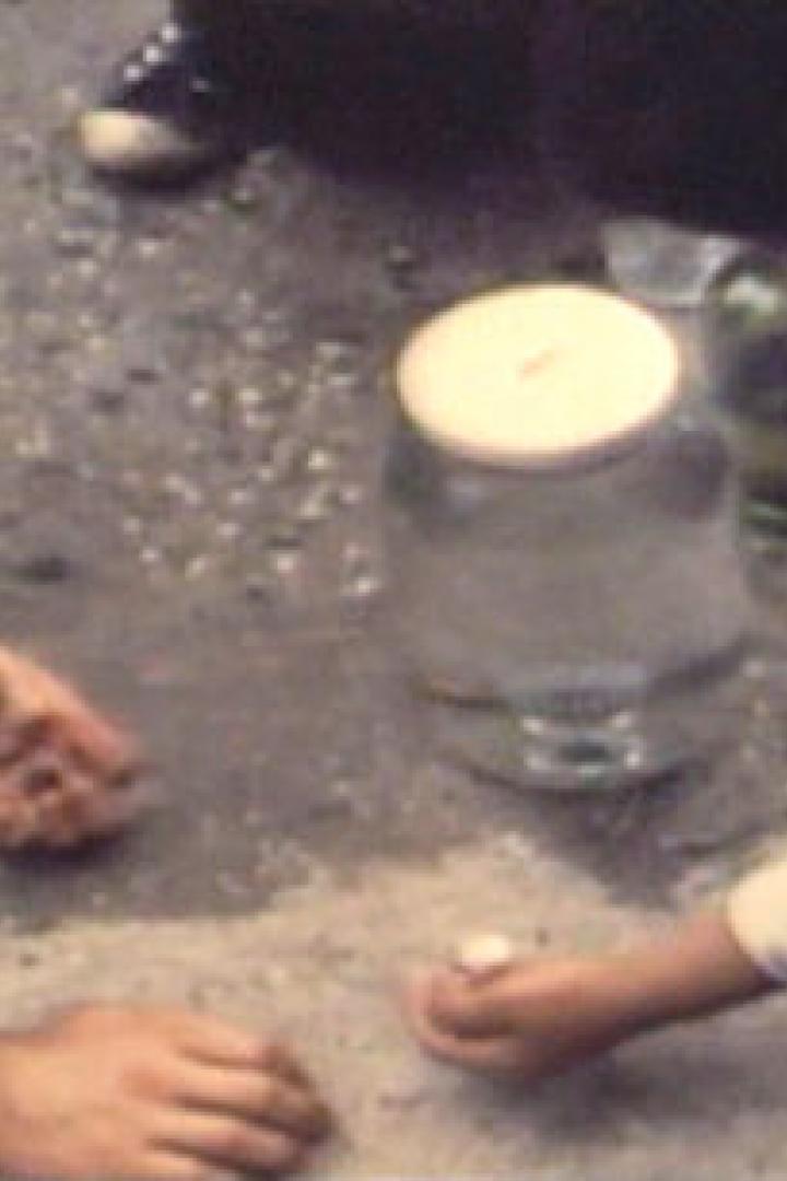Zwei Personen spielen Murmeln auf dem Boden, neben ihnen ein großes Glas mit Deckel.
