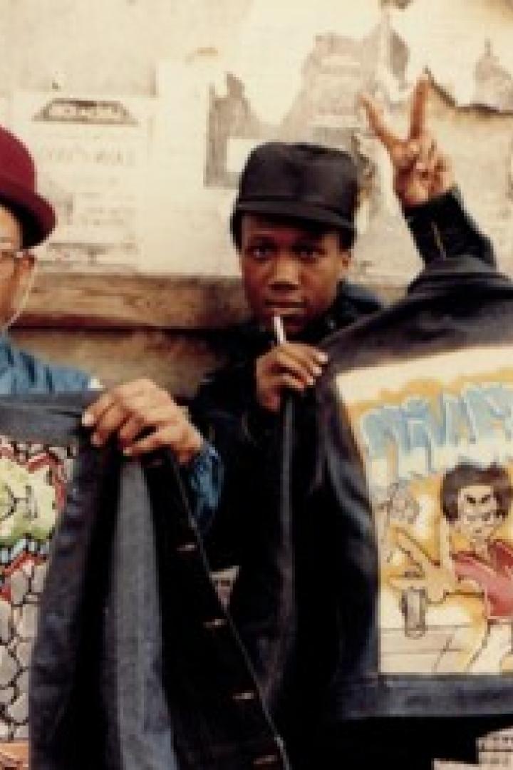 Drei Männer posieren mit bemalten Jeansjacken vor einer Wand mit Plakaten.