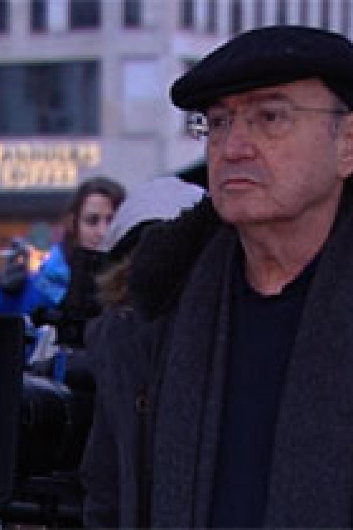 Ein älterer Mann mit Brille und Mütze steht vor einer Kamera, im Hintergrund ein Starbucks.