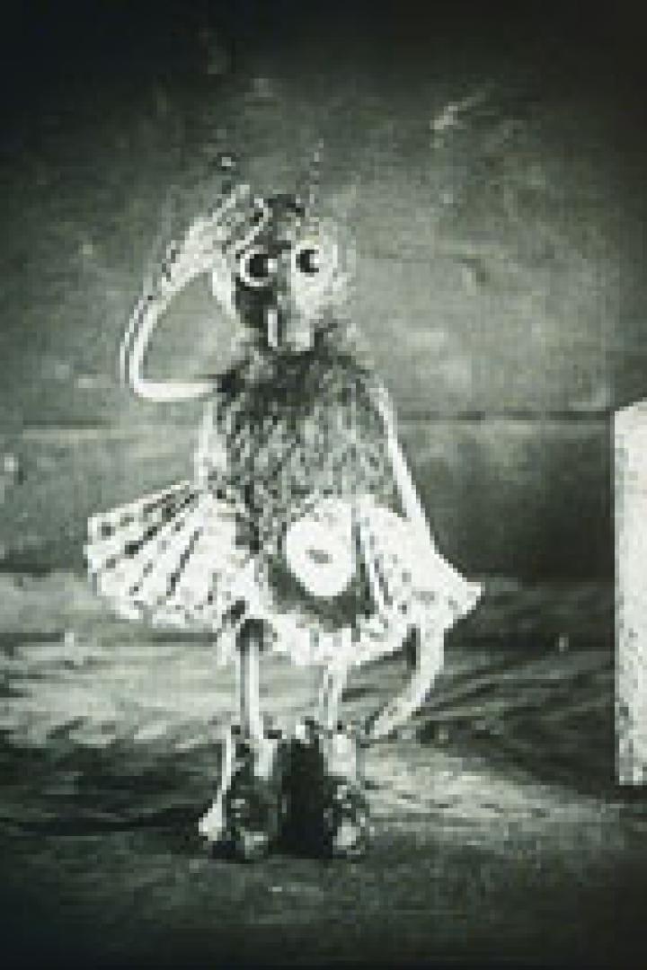 Eine anthropomorphe Figur mit Tutu und großen Augen steht vor einer Kiste.