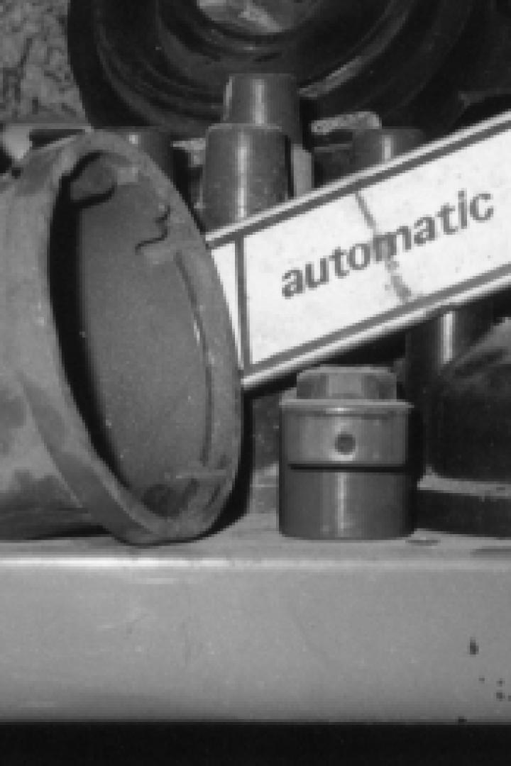 Verschiedene Autoteile liegen auf einem Regal, darunter ein Zündverteiler und ein Schild mit der Aufschrift „automatic“.