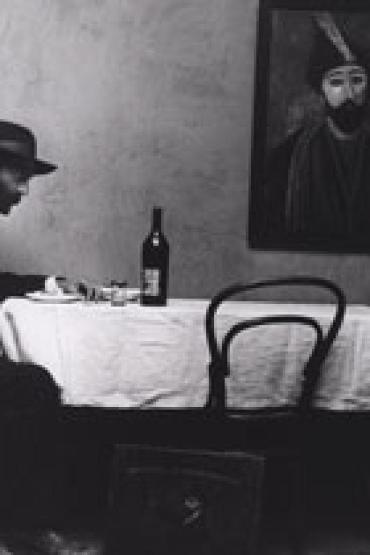 Ein Mann mit Hut sitzt an einem Tisch mit einer Flasche Wein und einem Gemälde an der Wand.