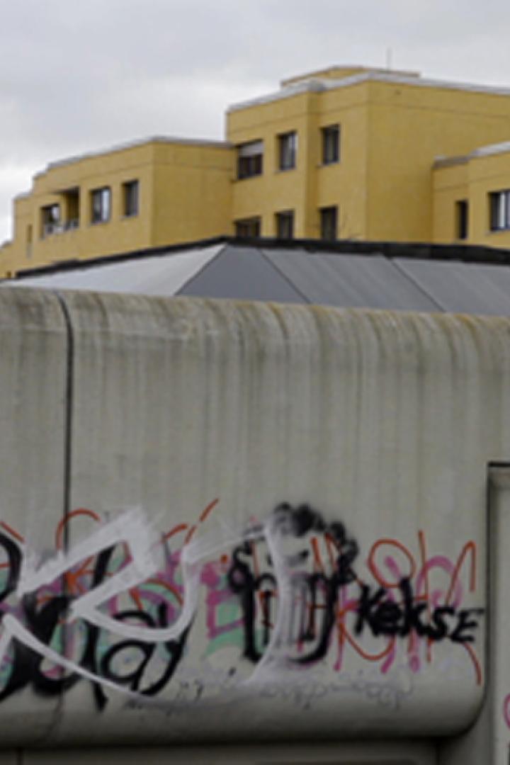 Eine Betonmauer mit Graffiti vor gelben Gebäuden unter einem grauen Himmel.