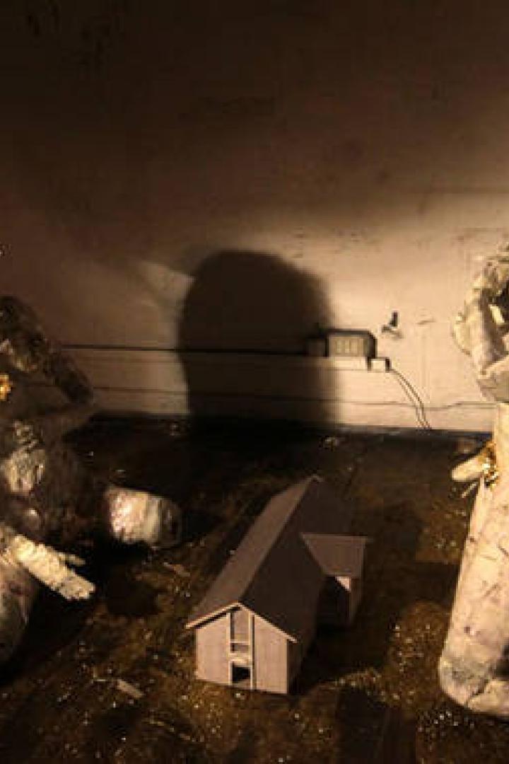 Zwei skulpturale Figuren mit goldenem Lametta knien vor einem kleinen Hausmodell in einem dunklen Raum.