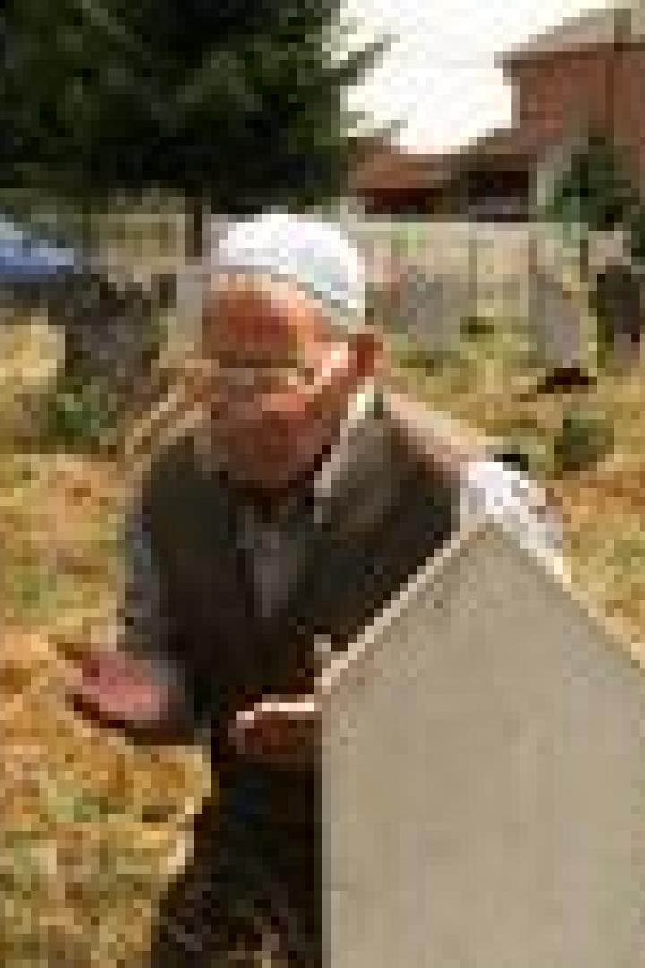 Ein älterer Mann mit Gebetskappe betet auf einem Friedhof vor zwei Grabsteinen.