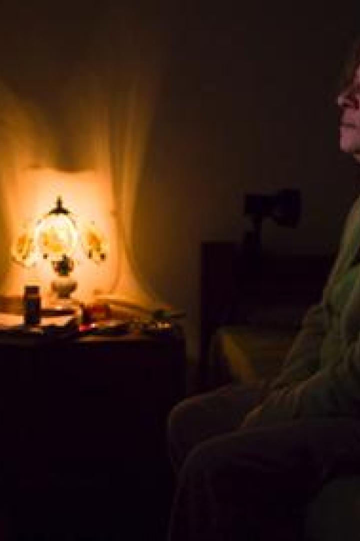 Eine Frau sitzt im Halbdunkel auf einem Bett und schaut nachdenklich zur Seite.