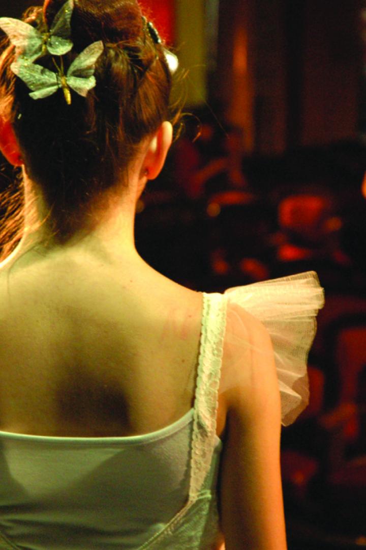 Eine Ballerina mit Schmetterlingen im Haar steht auf der Bühne vor leeren Sitzen.